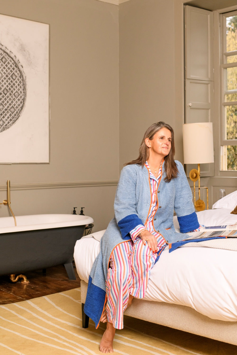 Model wearing Ramona blue long towelling robe with orange detail in the bedroom